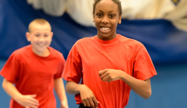 A schoolgirl smiles while jogging among fellow pupils in a sports hall.