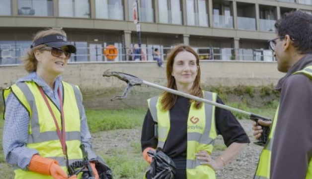 Three people talk while wearing high-vis vests and holding bags and litter-pickers.
