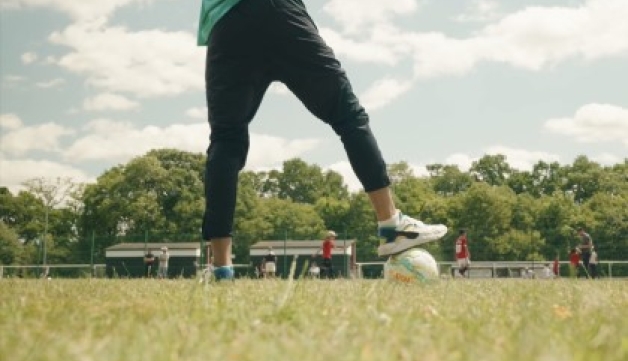 Close-up of a foot stood on top a football in the foreground of an amateur match, on a bright summer's day.