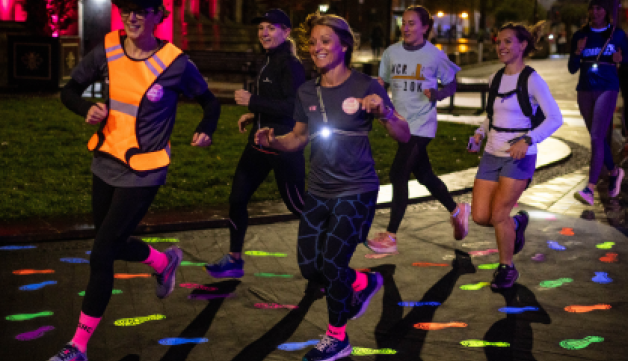 A group of women run at night down a road decorated with high-vis footprints.