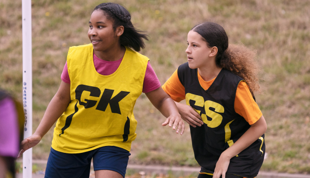 Two girls compete in a game of netball outdoors, wearing GK and GS vests.