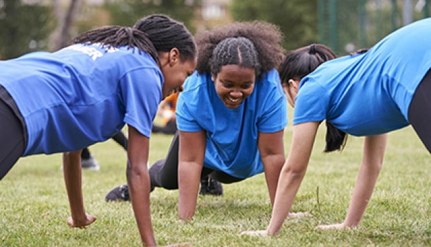 Three young girls adopt a press-up position on the grass outside, all facing each other in a circle