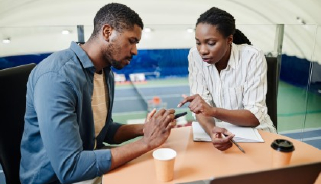 Two people consider something on a mobile phone while sat at a work desk above indoor tennis courts.