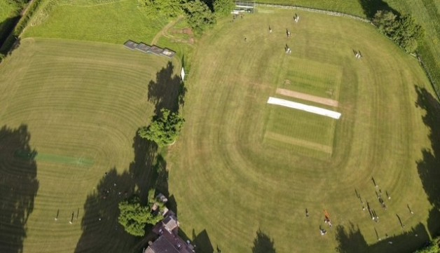 Aerial view of a game at Fillongley Cricket Club, showing the wicket, trees and clubhouse.