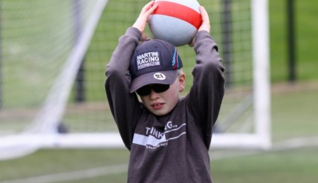 A boy wearing a baseball cap and dark glasses holds a ball above his head on an outdoor artificial football pitch.