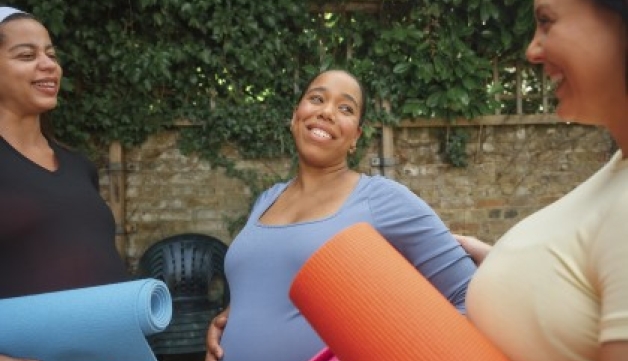 Three pregnant women stand talking outside while holding yoga mats.