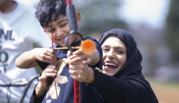A crouching mum helps her son hold his bow and arrow while he aims during an archery session.