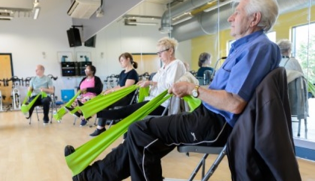 Several older adults use stretch their legs using resistance bands during a seated exercise class in a community hall.