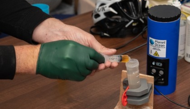 Close-up of a person's hands testing river water quality using a portable 'Planet Ocean' kit on a table.
