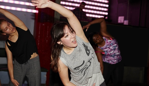 Women stretch their arms over their head during an exercise class in a studio.