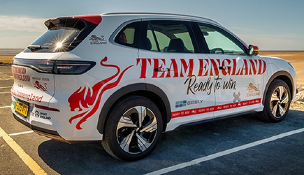 Branded Team England and Sport England electric taxi parked by a wide sandy beach on a sunny day under a clear blue sky.