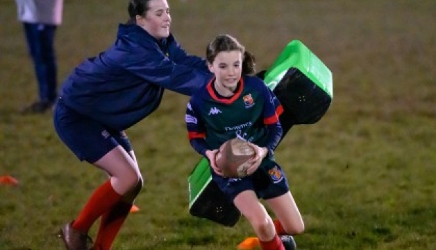 A young female rugby player runs with the ball and evades another girl holding a tackle pad during a training session at night.