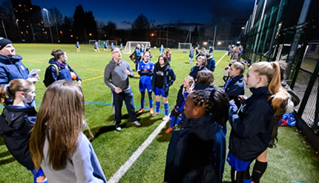 A coach addresses a girls football team during a session on an outdoors pitch.