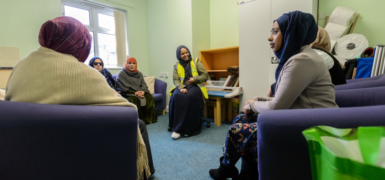 A group of women, several wearing headscarves and shawls, talk while sat in a group in a room.