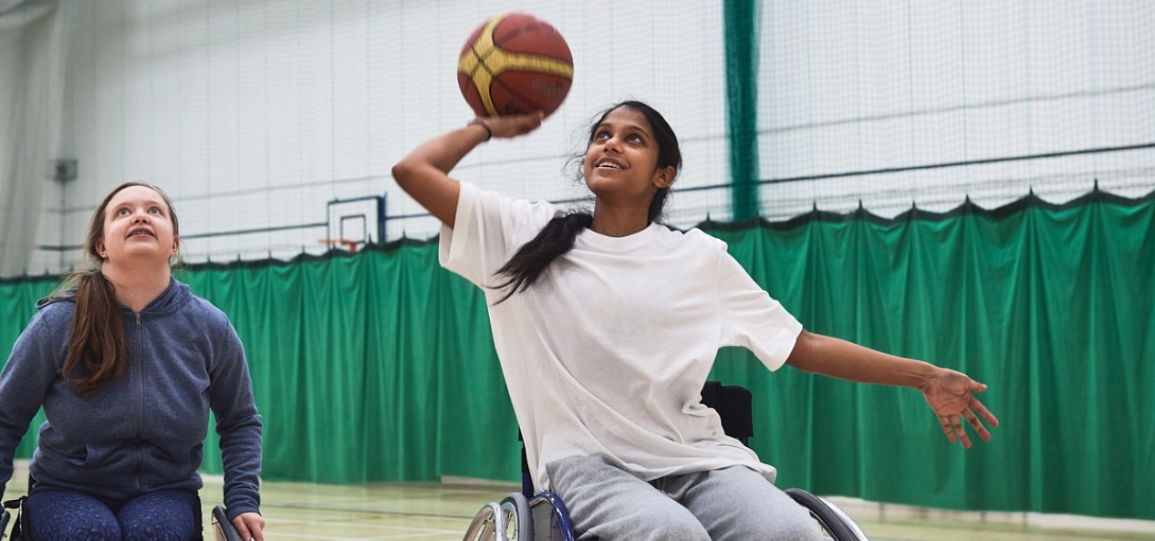 A young female wheelchair user prepares to shoot a basketball as a fellow player watches on.
