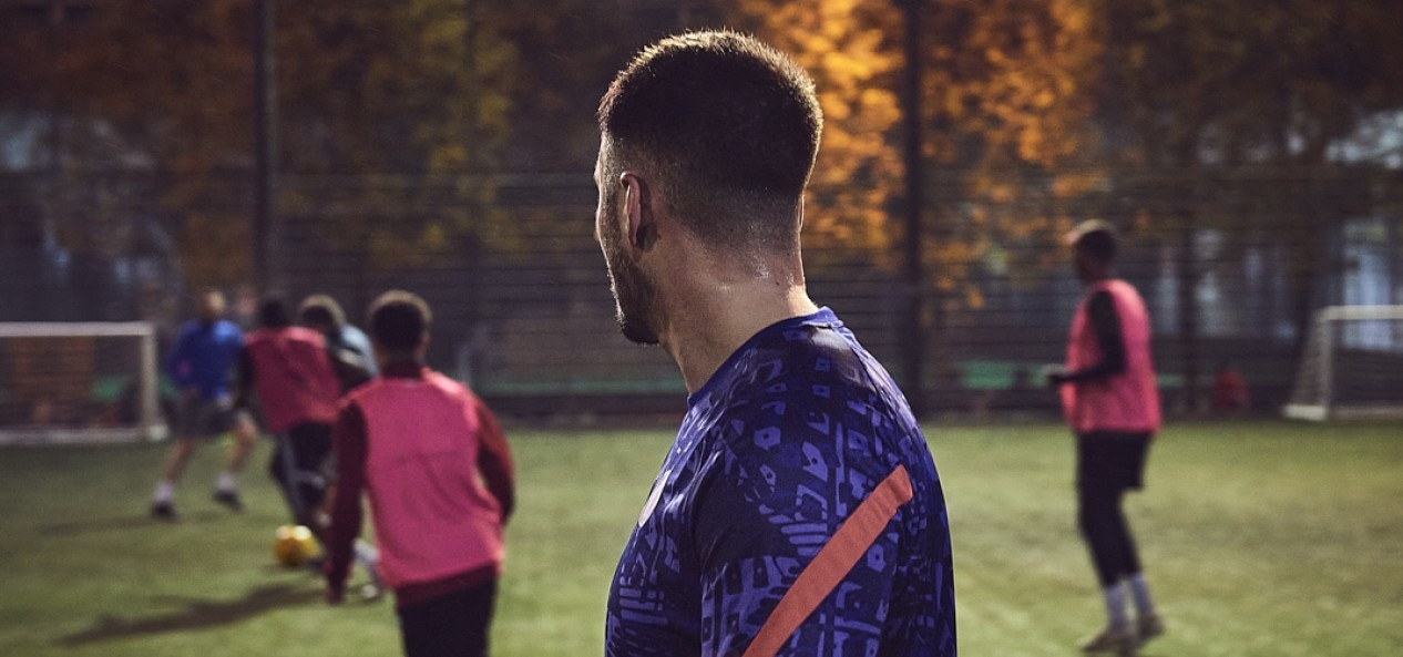 Shot from behind a casual footballer as he looks at his fellow players in action at night.