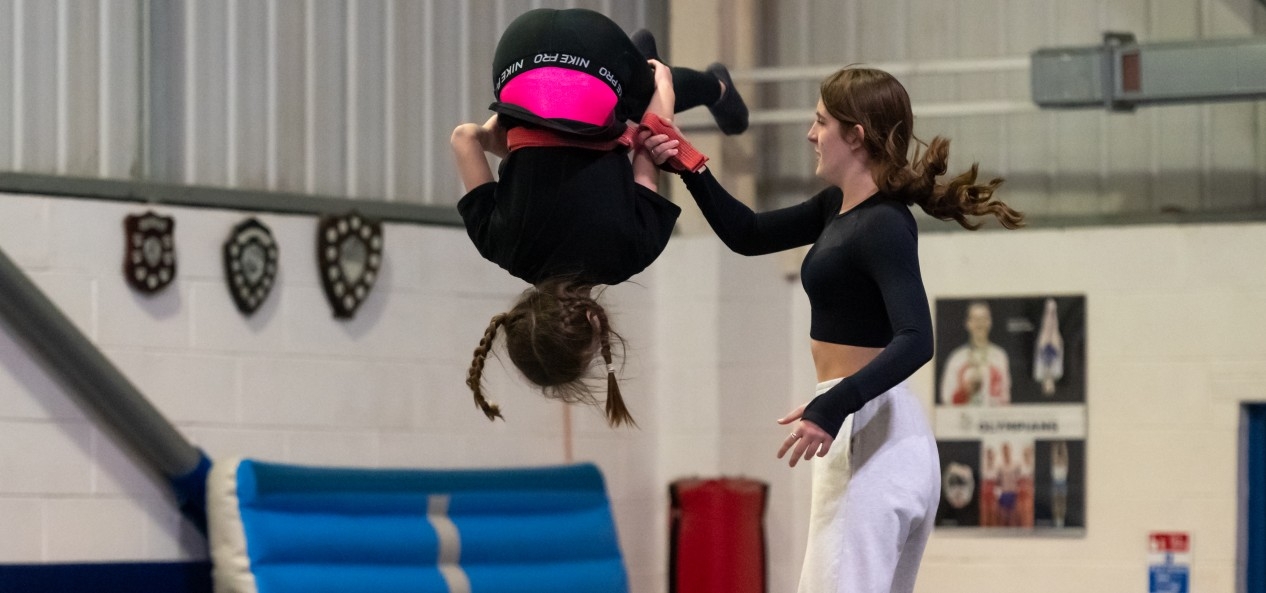 A female coach helps a young girl tumble in a gymnastics and trampolining hall.