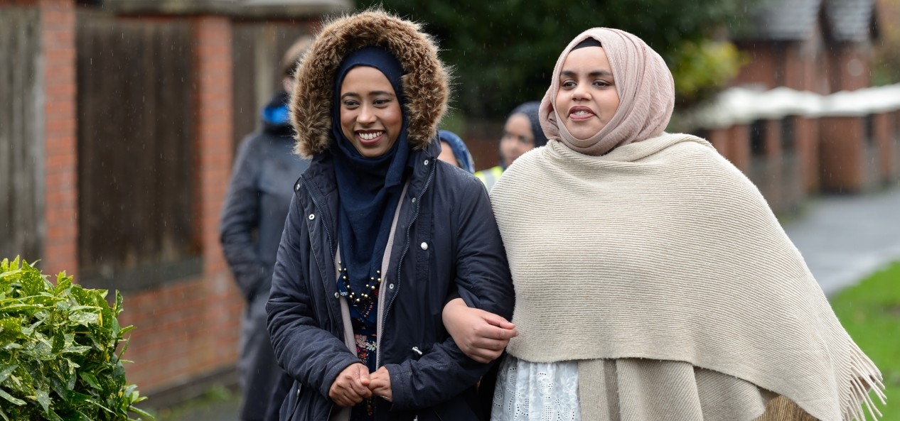 Two women wearing head shawls and coats link arms as they walk down a street.