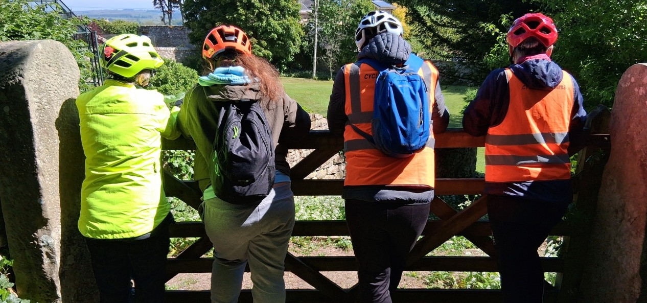 Four cyclists wearing helmets, hi-vis clothing and backpacks look over a fence into a field.