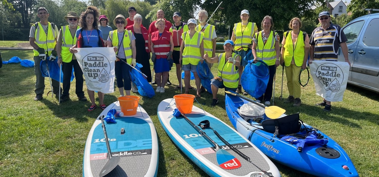 A group of people in high-vis vests pose for a photo, in front of paddle boards and a canoe, holding litter pickers and big bags