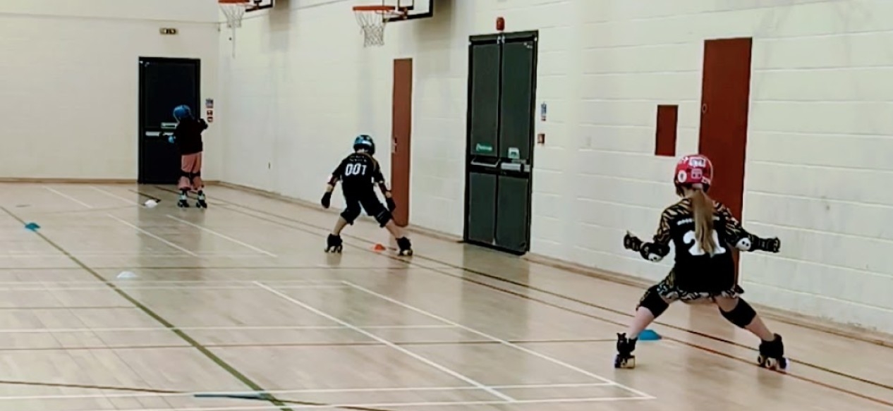 Three young people wearing helmets take part in a roller derby session in a sports hall.