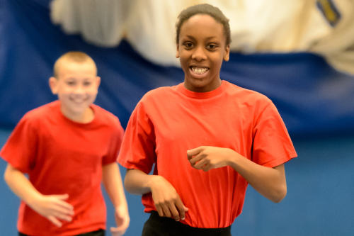 A schoolgirl smiles while jogging among fellow pupils in a sports hall.