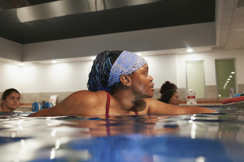 A woman wearing a bandana in a swimming pool reaches out to the side of the pool during an aqua aerobics class
