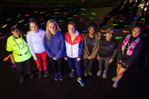 Seven women stand side by side looking upwards at the camera, with glow-in-the-dark footsteps in the background.
