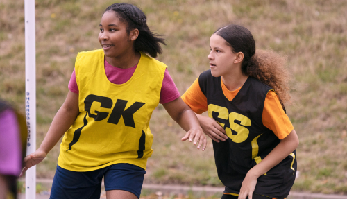 Two girls compete in a game of netball outdoors, wearing GK and GS vests.