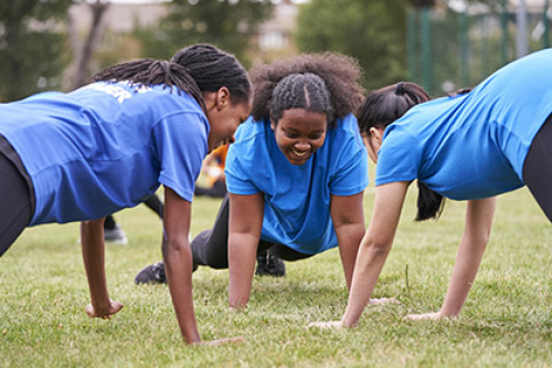 Three young girls adopt a press-up position on the grass outside, all facing each other in a circle