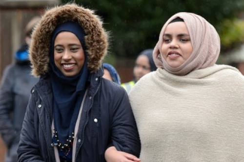 Two women, one with her coat's hood up and the other wearing a headscarf and shawl, link arms as they walk side by side.