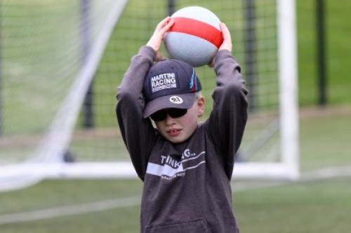 A boy wearing a baseball cap and dark glasses holds a ball above his head on an outdoor artificial football pitch.