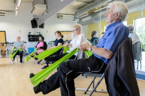Several older adults use stretch their legs using resistance bands during a seated exercise class in a community hall.