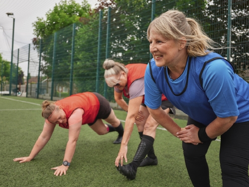 A group of women perform stretches on an artificial grass pitch, ahead of a walking football match