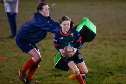 A young female rugby player runs with the ball and evades another girl holding a tackle pad during a training session at night.