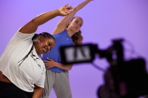Two Studio You x Nike trainers stretch one arm over their heads as a camera in the foreground films them.