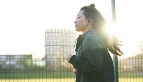 A young woman wearing earphones jogs by a fence in an urban park, with the sun glinting behind her..