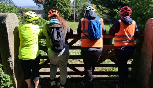 Four cyclists wearing helmets, hi-vis clothing and backpacks look over a fence into a field.