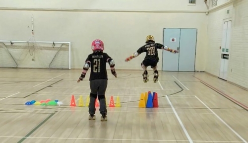 Two young people wearing helmets take part in a roller derby session, jumping over cones in a sports hall.