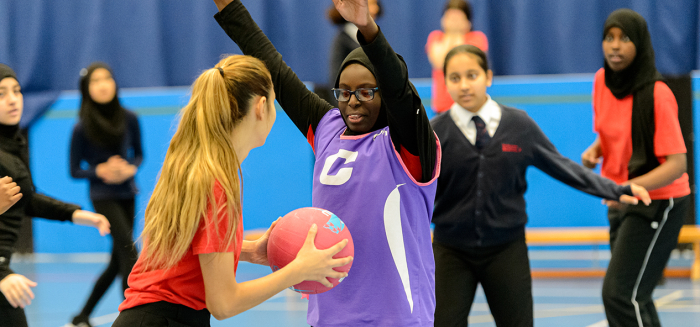 Girls play netball in a PE lesson. One player has the ball and another girl is guarding her, with both arms in the air.