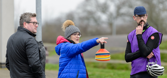 A woman holding a stack of marker cones points while talking to two men, one of whom is holding a cycling helmet.
