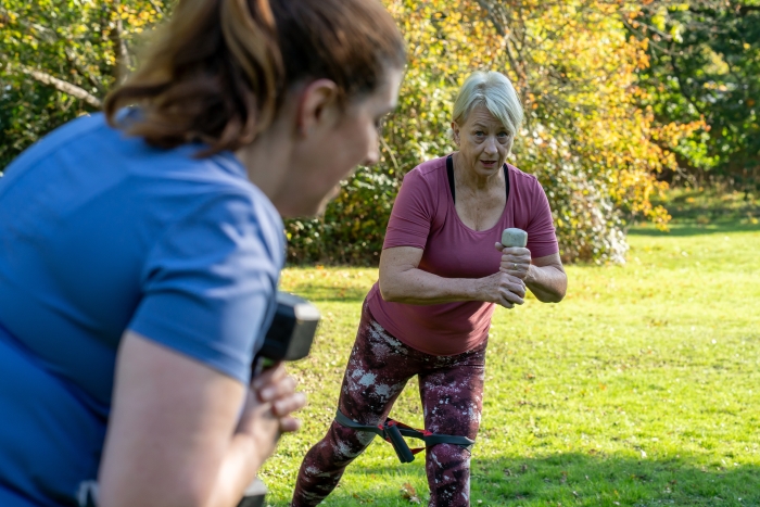 Two women exercise outdoors in a park on a sunny day, each holding a dumbbell on the grass with trees in the background.