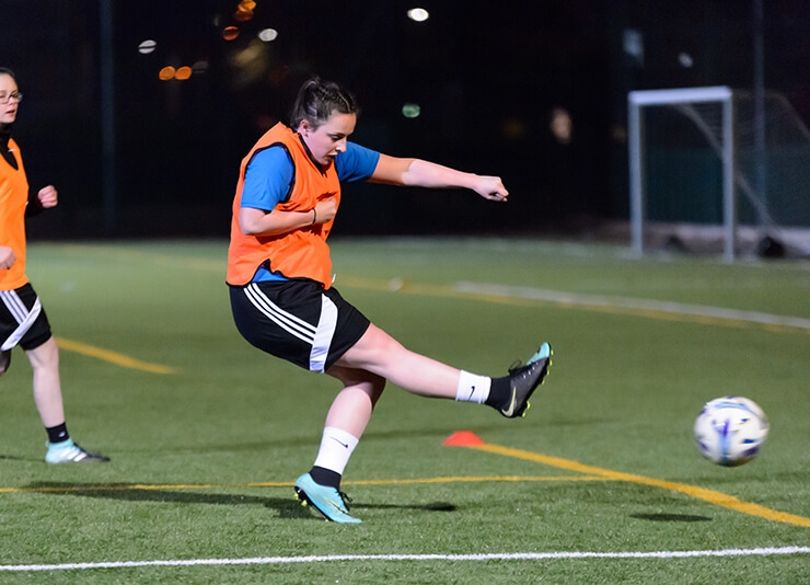 A woman kicking a football while playing on an outdoors pitch.