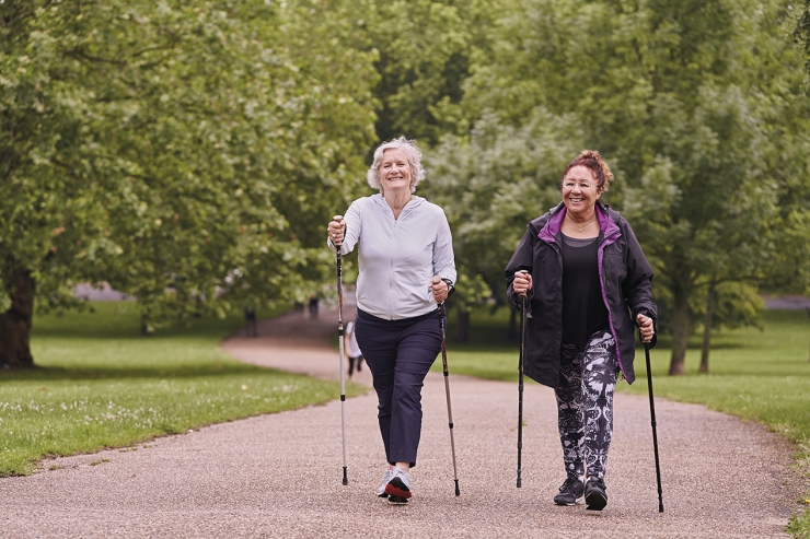 Two women walk with poles on a path in a park.