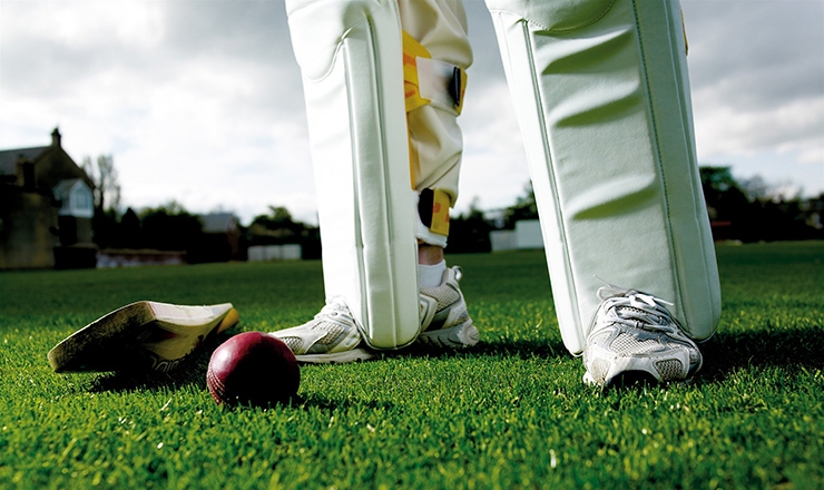 A cricket bat and ball on the grass with a cricketer's legs in the background