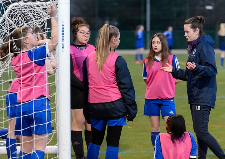A female coach speaks to a teenage girls' football team at the side of an artificial football pitch