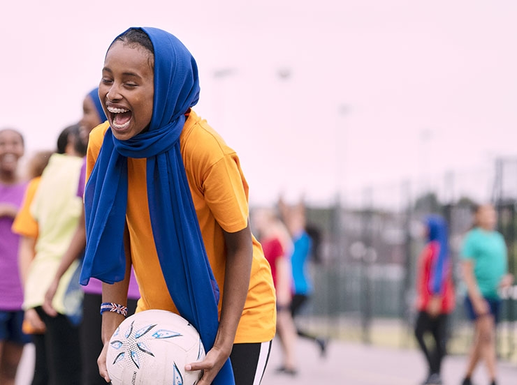 A girl wearing a head scarf laughs and smiles while playing netball in a school playground