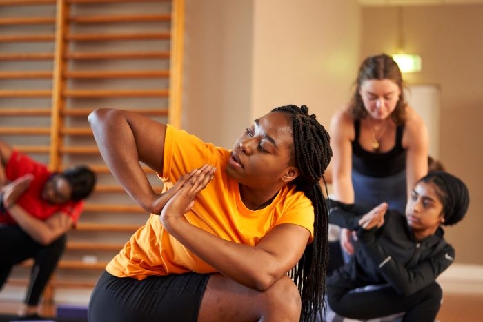 Young women are taking part in an exercise class in a hall, their hands are clasped as though in prayer