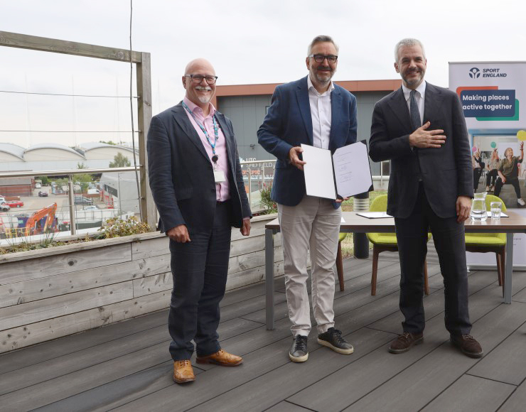 Sport England chief executive Tim Hollingsworth, South Yorkshire mayor Oliver Coppard, and South Yorkshire Integrated Care Board CEO Gavin Boyle sign a memorandum of understanding at a table outside.