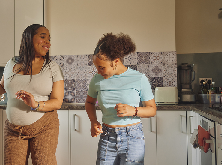 Two women, the one on the left being pregnant, dance in a kitchen